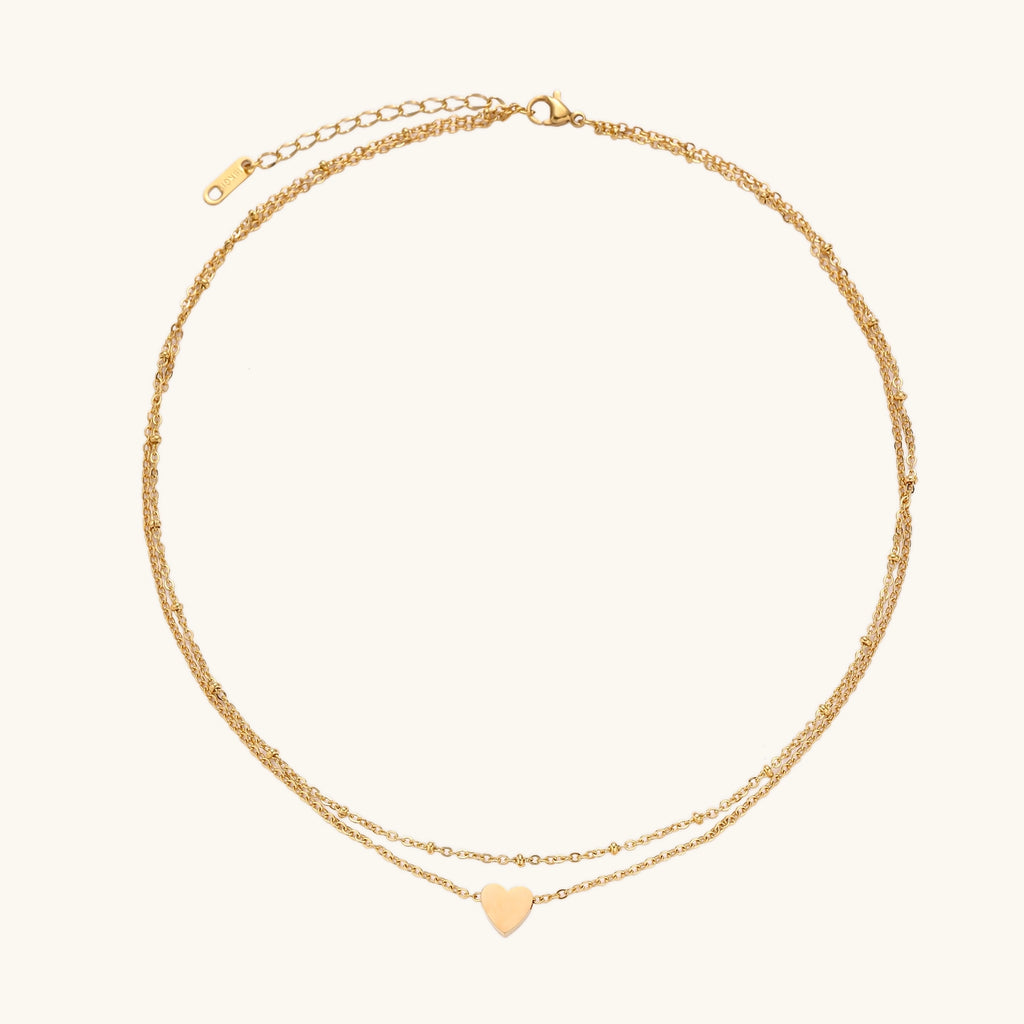 Two-layer gold-plated stainless steel necklace with small heart pendant displayed on beige background
